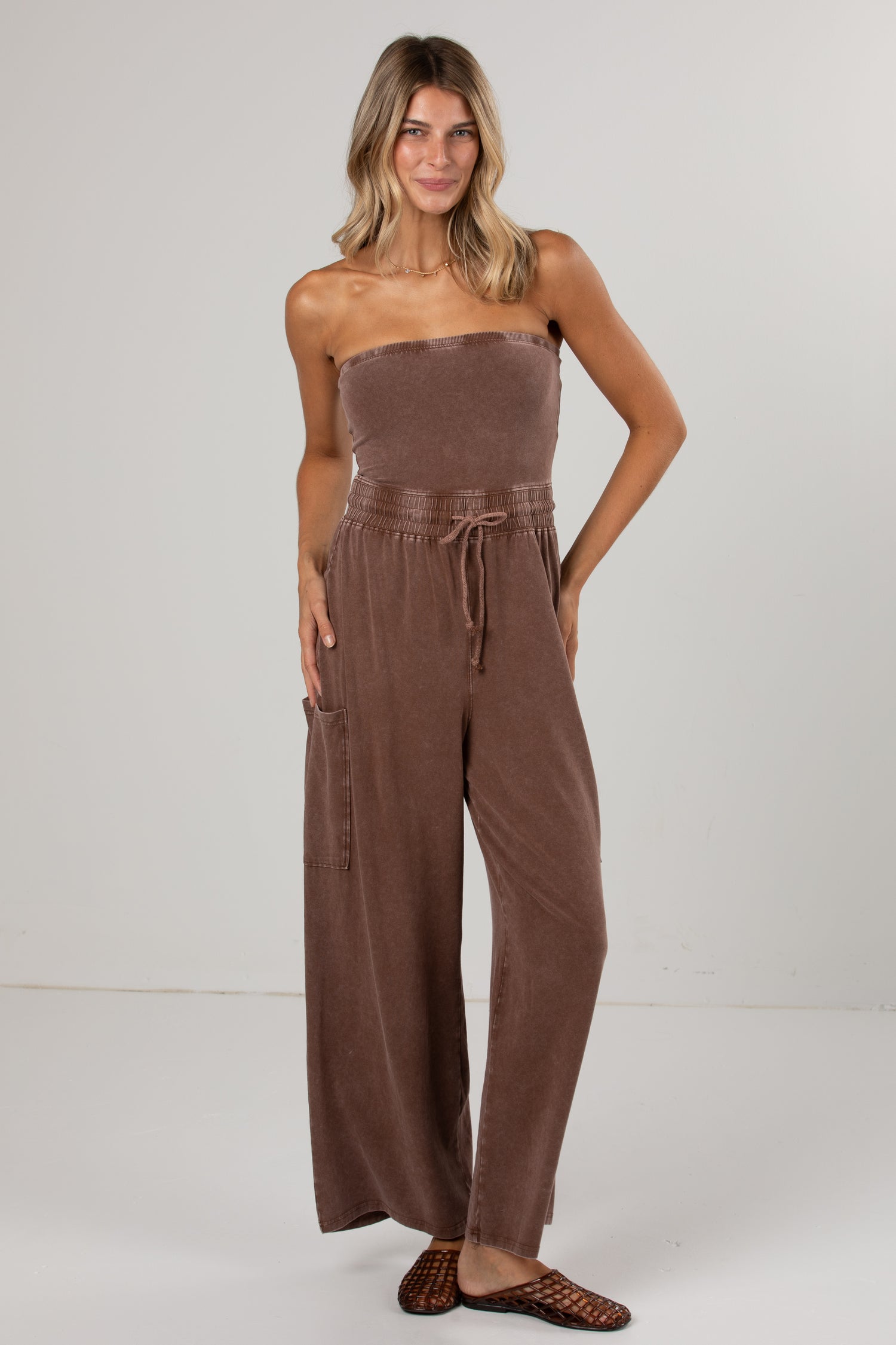 A woman modeling a brown strapless wide-leg jumpsuit with a drawstring waist and side pockets, paired with brown woven flat shoes, against a light gray studio background.