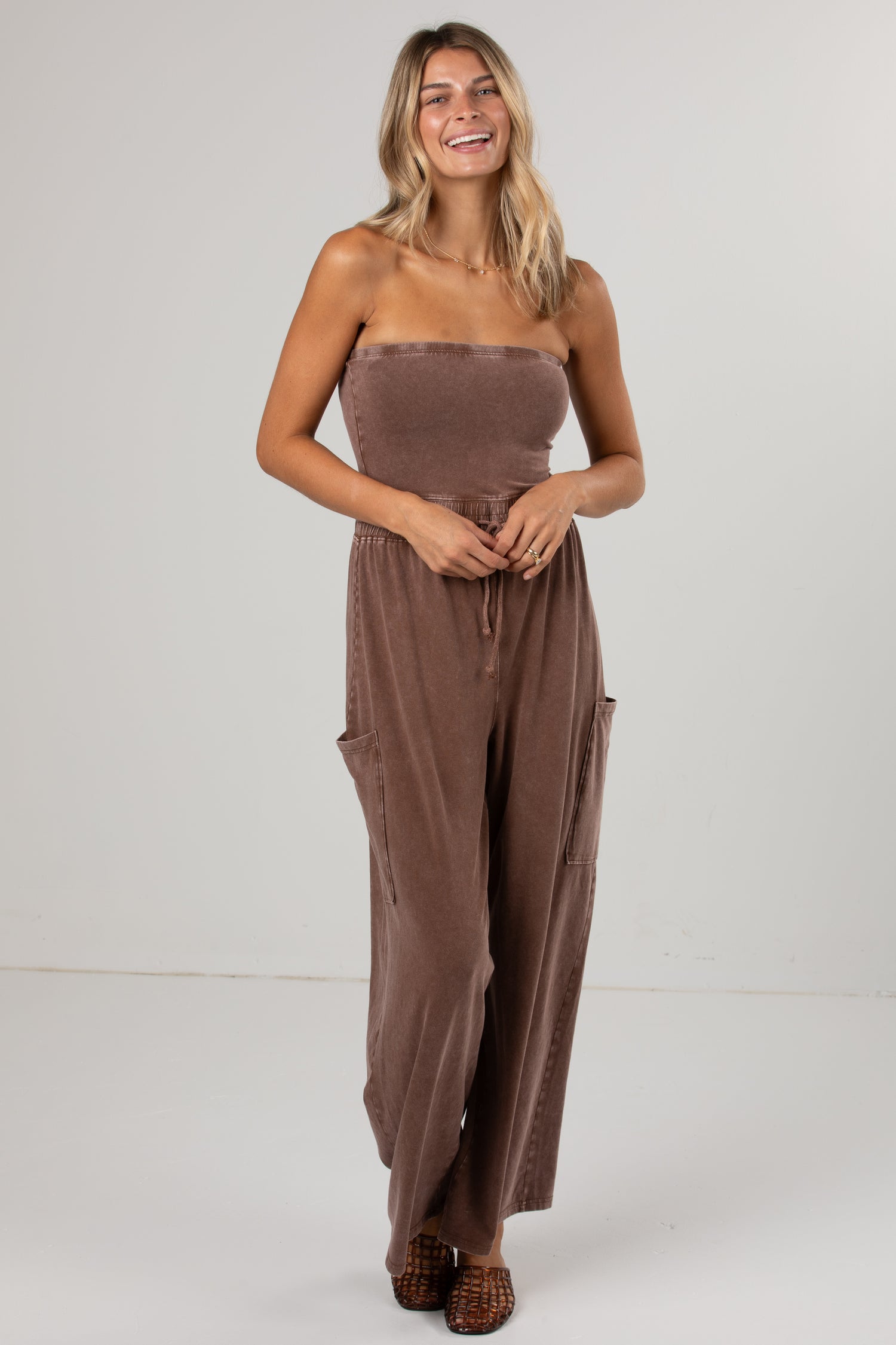 A woman modeling a brown strapless jumpsuit with wide legs and side cargo pockets, standing against a plain light gray studio background.
