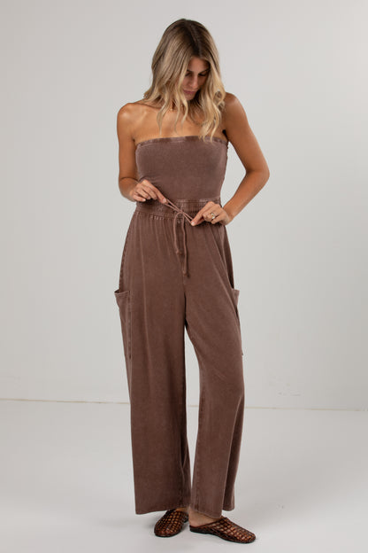 A woman modeling a brown strapless wide-leg jumpsuit with a drawstring waist and side pockets, paired with brown woven flat shoes, against a plain light gray studio background.
