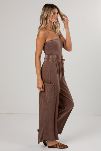 A woman modeling a strapless brown wide-leg jumpsuit with a matching drawstring waist belt, photographed against a plain light gray studio background.