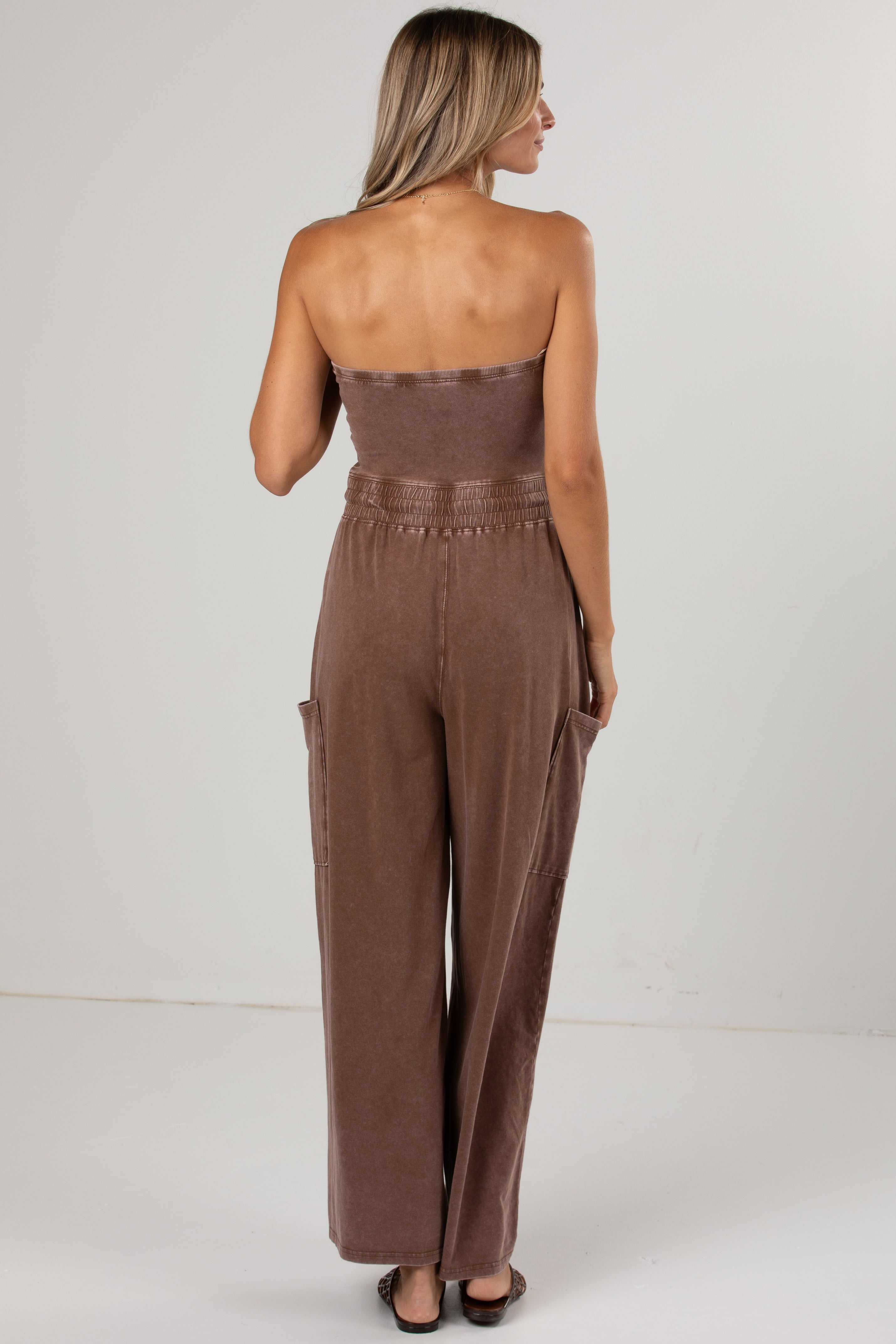 A woman shown from the back wearing a brown strapless jumpsuit with wide legs and side pockets, standing against a plain light gray studio background.
