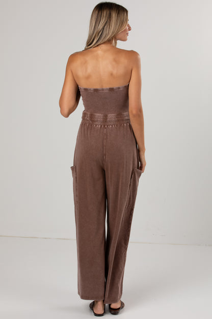 A woman shown from the back wearing a brown strapless jumpsuit with wide legs and side pockets, standing against a plain light gray studio background.