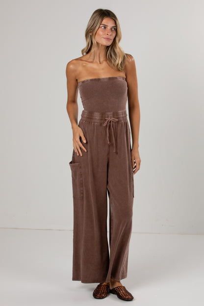 A woman modeling a brown strapless wide-leg jumpsuit with a drawstring waist and side pockets, standing against a plain light gray studio background.