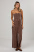 A woman modeling a brown strapless wide-leg jumpsuit with a drawstring waist and side pockets, standing against a plain light gray studio background.