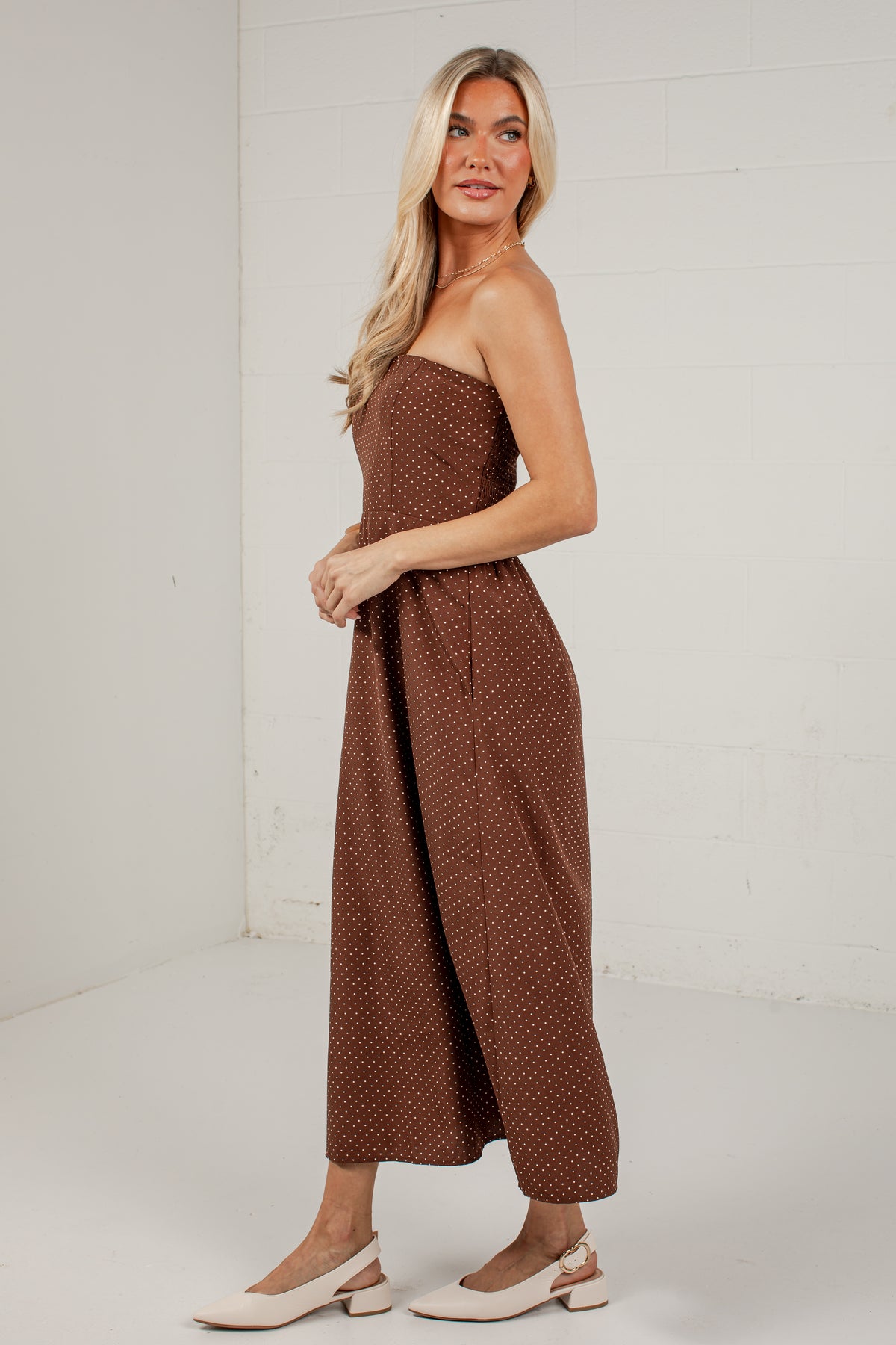 A woman modeling a brown strapless jumpsuit with a small white polka dot pattern, paired with white Mary Jane shoes, standing in a white studio corner.