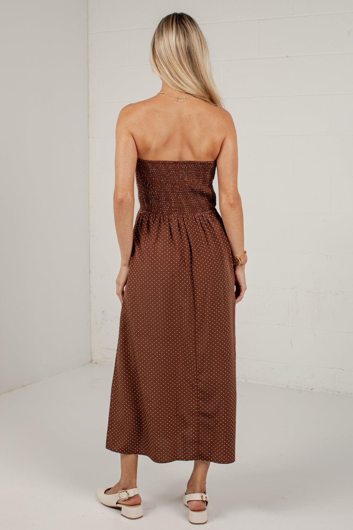 A woman viewed from behind wearing a strapless brown midi dress with a smocked bodice and small white polka dots, paired with white platform sandals, set against a white brick wall.