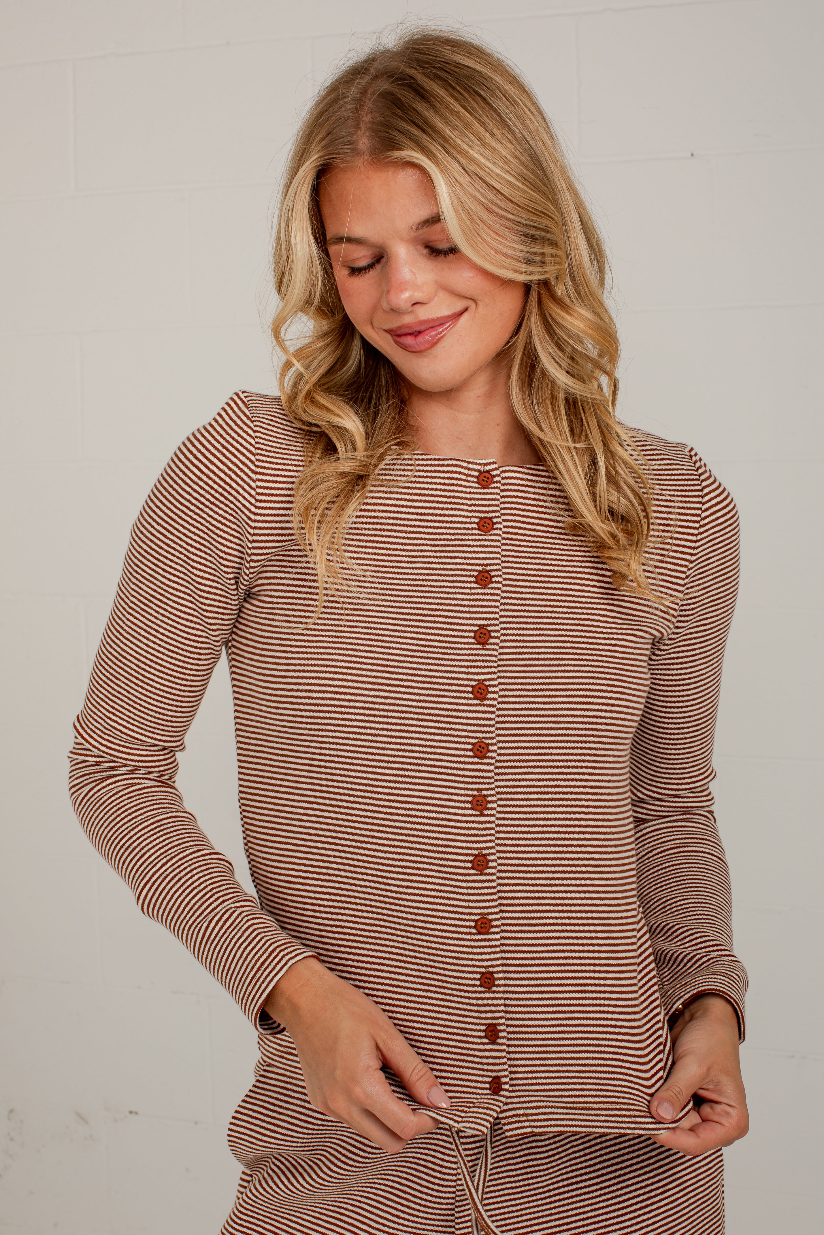 A woman wearing a long-sleeve button-front top with thin horizontal brown and beige stripes, standing against a light neutral wall.