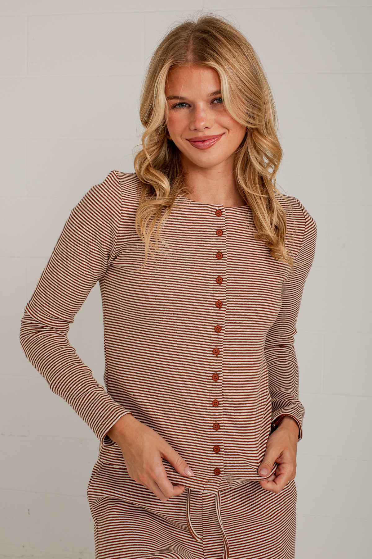 A woman modeling a long-sleeve knit top with thin brown and beige horizontal stripes, featuring a button-down front and a drawstring waist, set against a light gray brick wall.