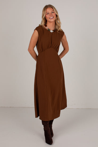 A woman modeling a sleeveless brown midi dress with a gathered waist and pleated skirt, paired with dark brown ankle boots, against a plain light gray studio background.