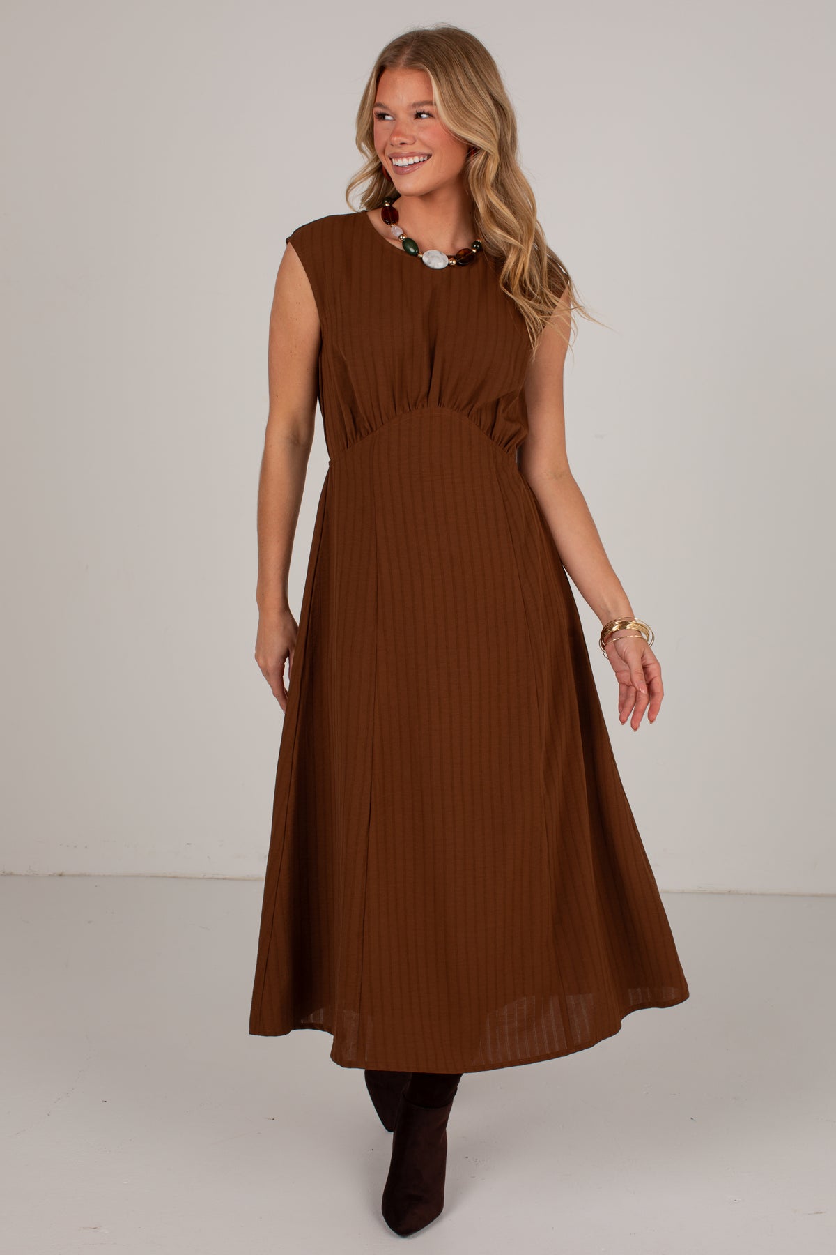 A woman modeling a sleeveless brown midi dress with a gathered waist and pleated skirt, standing against a plain light gray studio background.