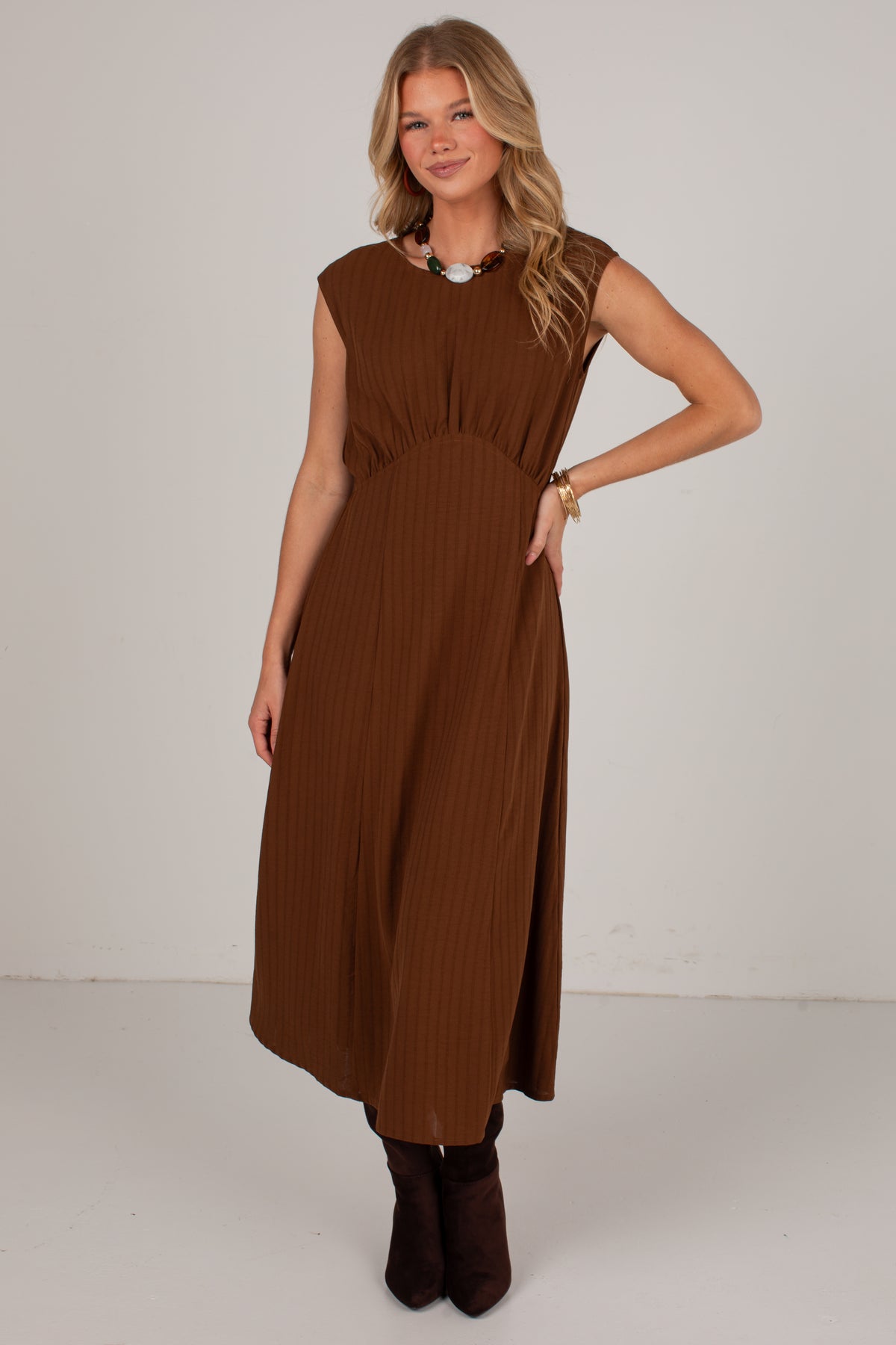A woman modeling a sleeveless brown midi dress with a gathered waist and pleated texture, paired with dark brown ankle boots against a plain light gray studio background.