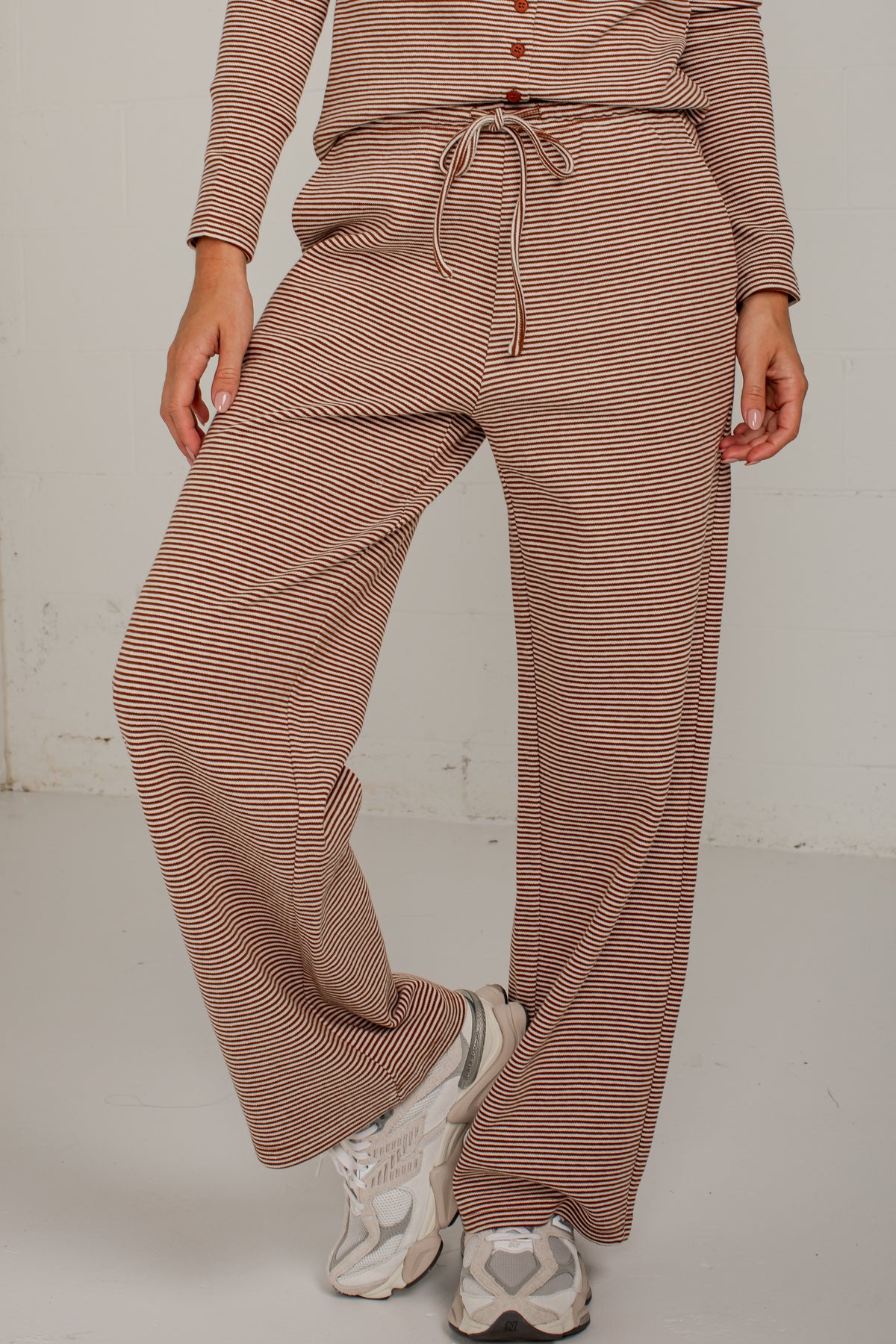 A person wearing loose, wide-leg knit pants with thin brown and beige horizontal stripes, a matching drawstring waist, and a matching long-sleeve top, paired with white sneakers against a plain light gray studio background.