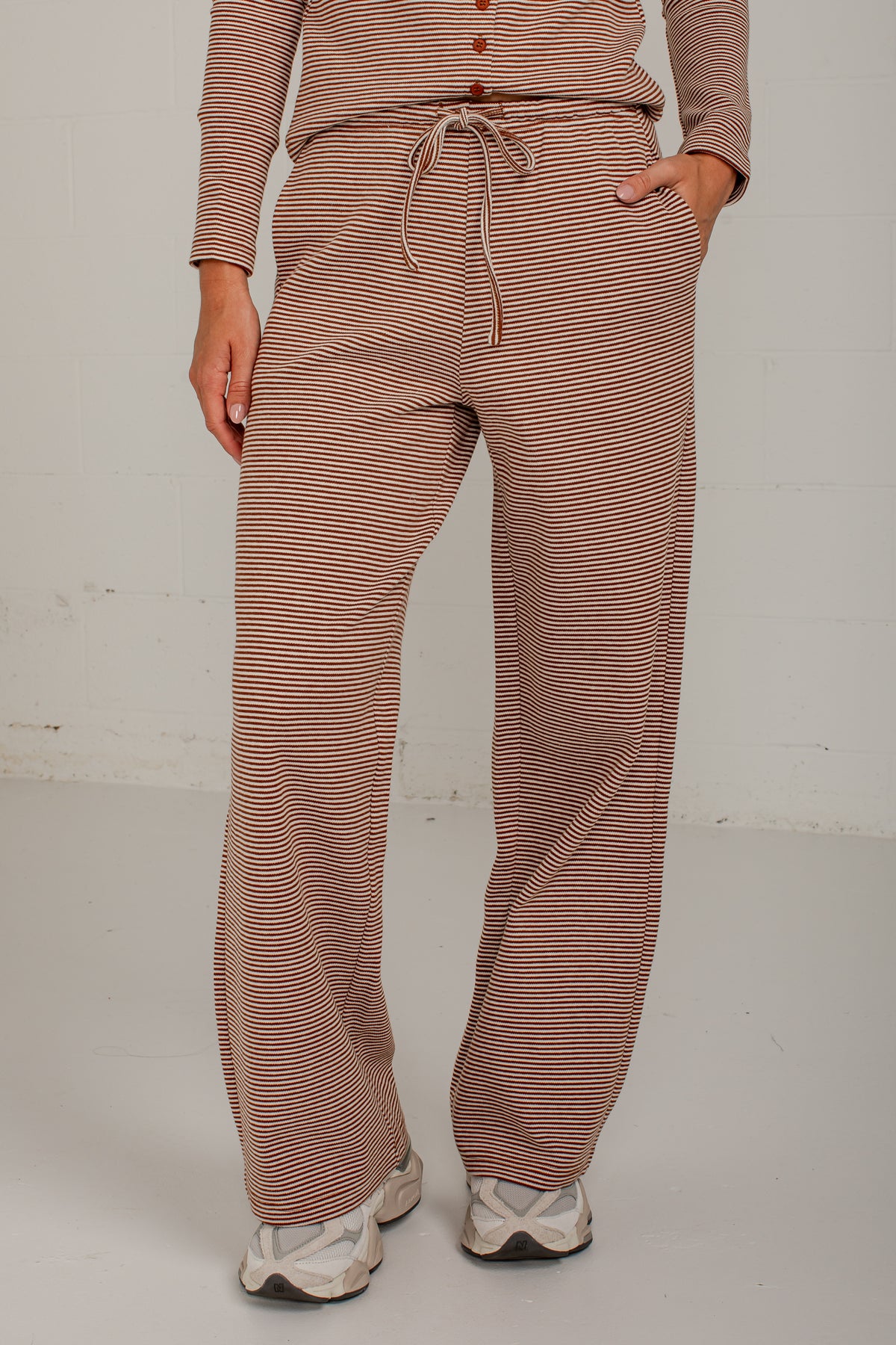 A person wearing loose, wide-leg drawstring pants with thin horizontal brown and beige stripes, paired with a matching long-sleeve top and white sneakers, standing against a light gray wall.