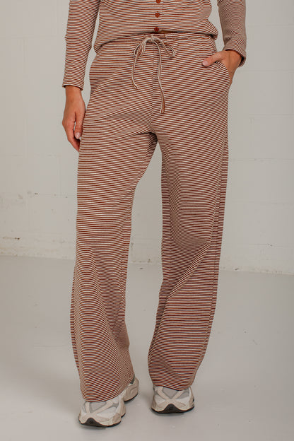 A person wearing loose, wide-leg drawstring pants with thin horizontal brown and beige stripes, paired with a matching long-sleeve top and white sneakers, standing against a light gray wall.