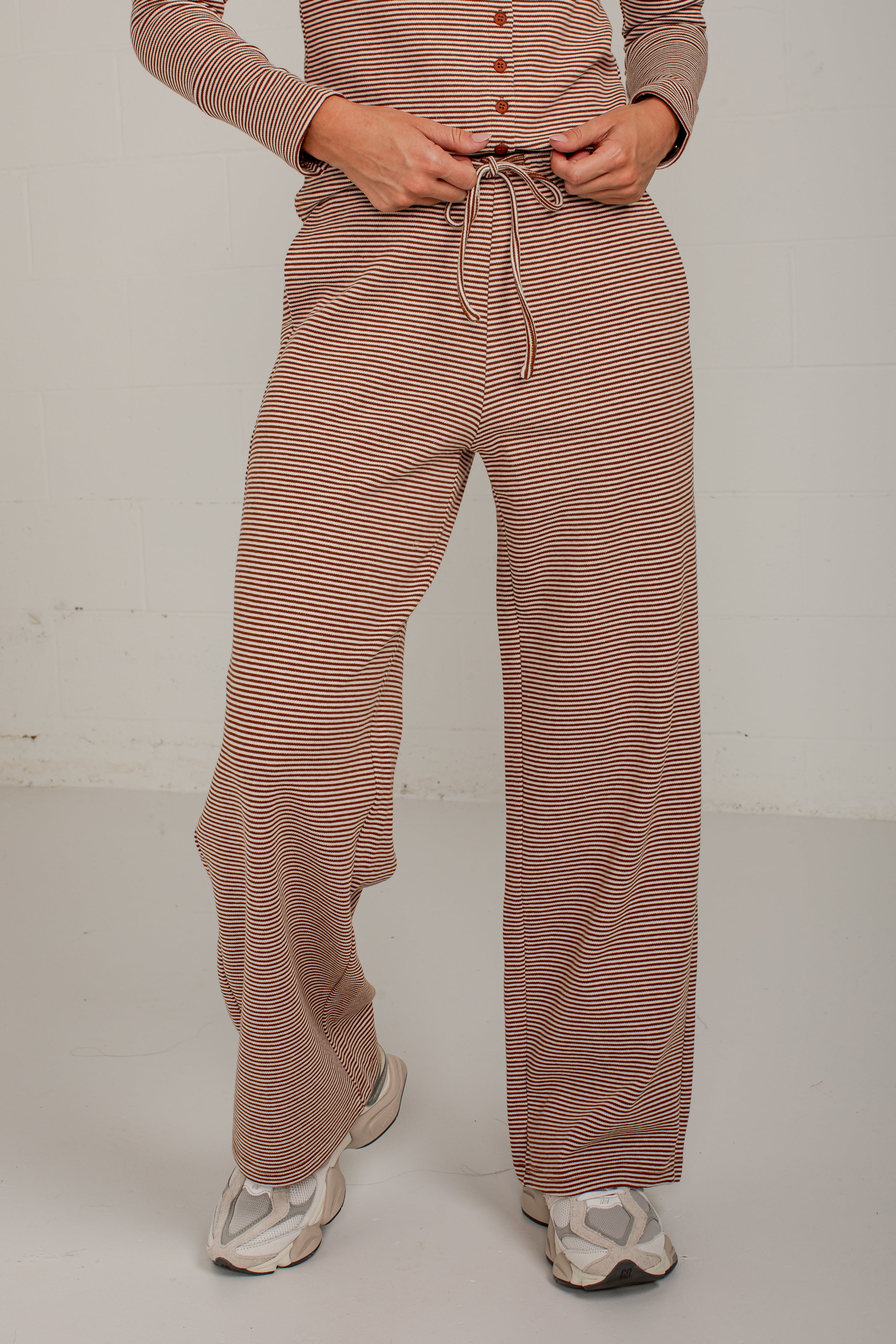 A person wearing loose, wide-leg knit pants with thin brown and beige horizontal stripes, paired with a matching long-sleeve top and white sneakers, standing against a plain light gray wall.