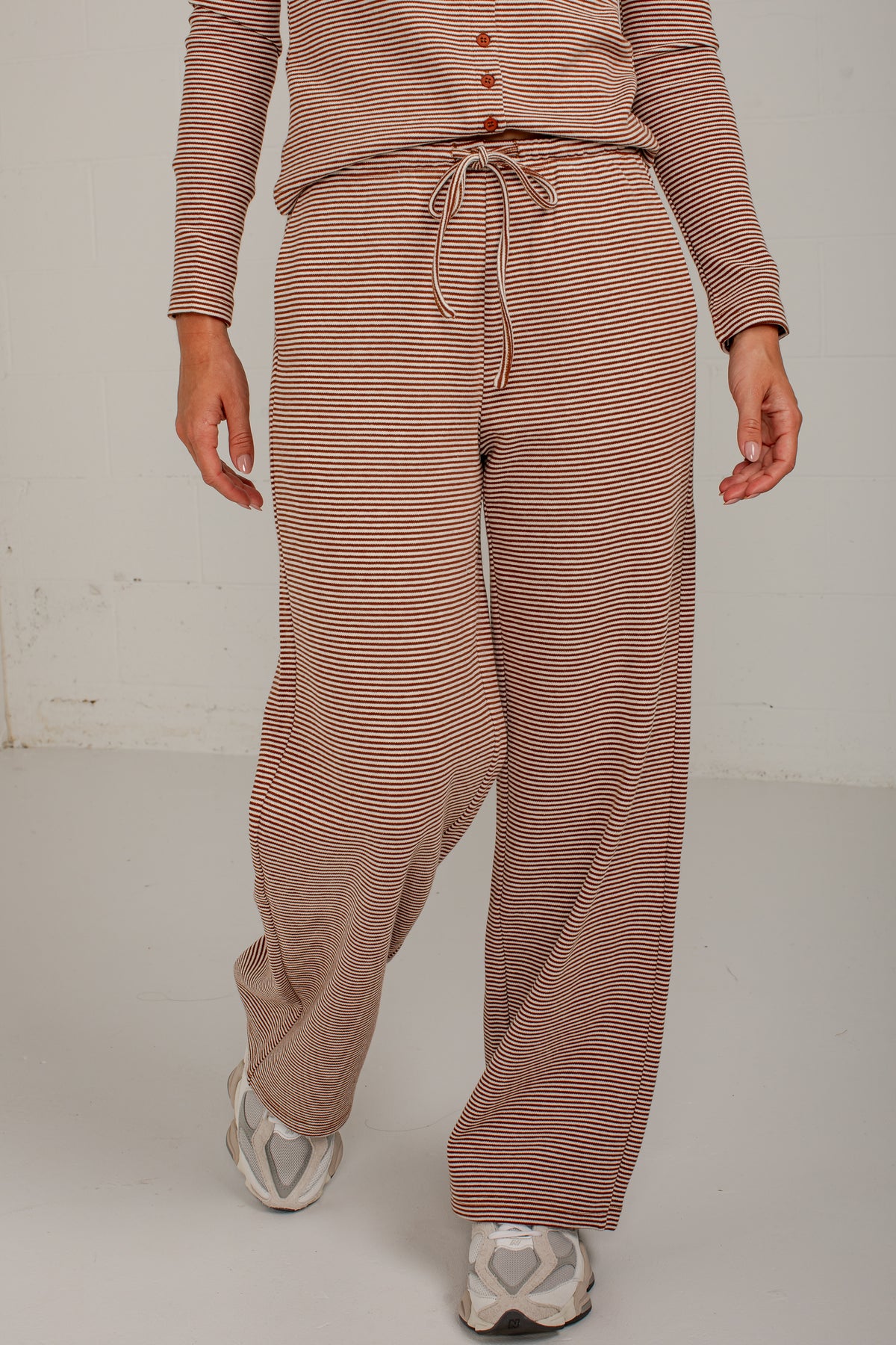 A person wearing loose, wide-leg knit pants and a matching long-sleeve top with thin horizontal stripes in a dusty rose and beige colorway, paired with white sneakers, against a plain off-white studio background.
