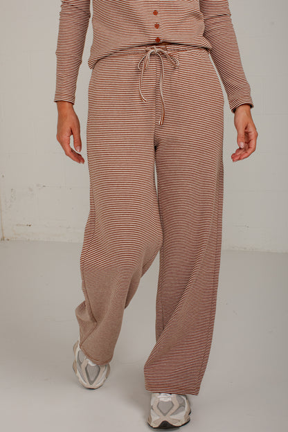 A person wearing loose, wide-leg knit pants and a matching long-sleeve top with thin horizontal stripes in a dusty rose and beige colorway, paired with white sneakers, against a plain off-white studio background.