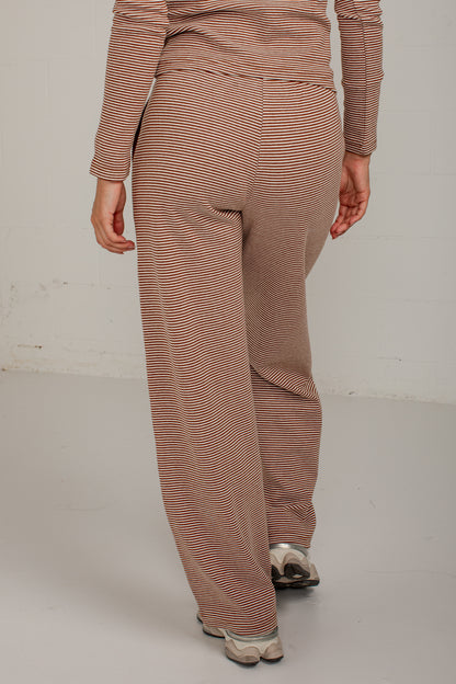 A person shown from the back wearing loose, wide-leg knit pants and a matching long-sleeve top with fine horizontal stripes in warm taupe and brown, standing against a plain light wall.