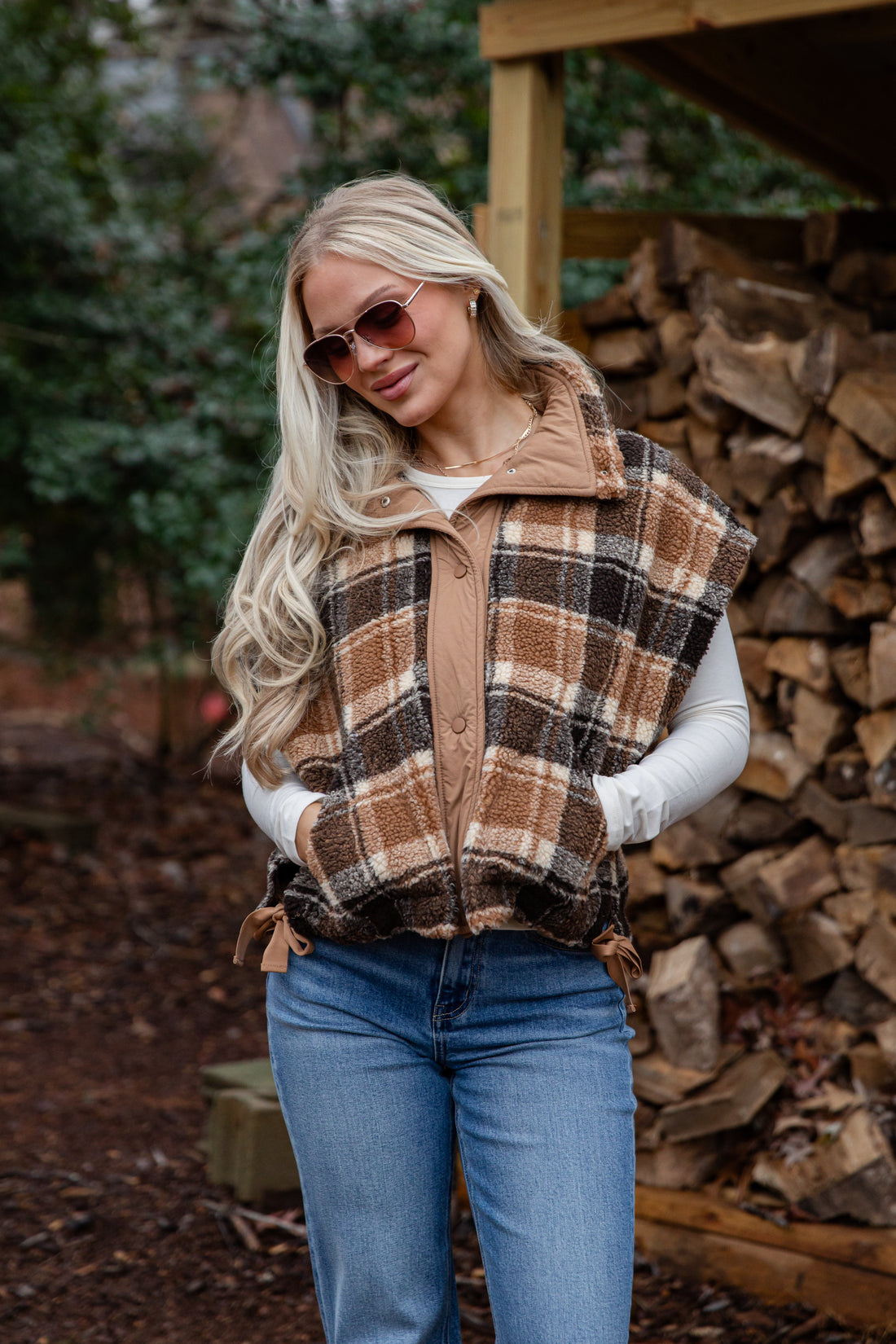 Woman wearing a plaid vest and sunglasses outdoors with stacked firewood in the background