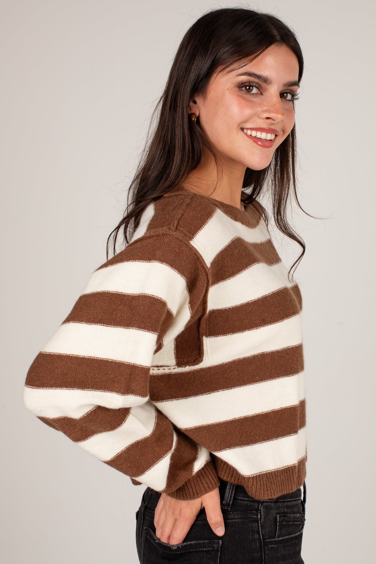 A woman wearing a brown and cream striped knit sweater, shown from a side-back angle against a plain light gray studio background.