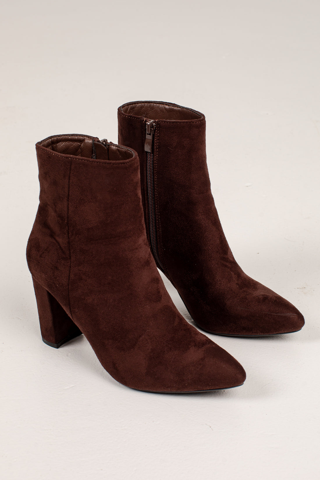 A pair of dark brown suede ankle boots with pointed toes and block heels, shown on a light gray studio background.