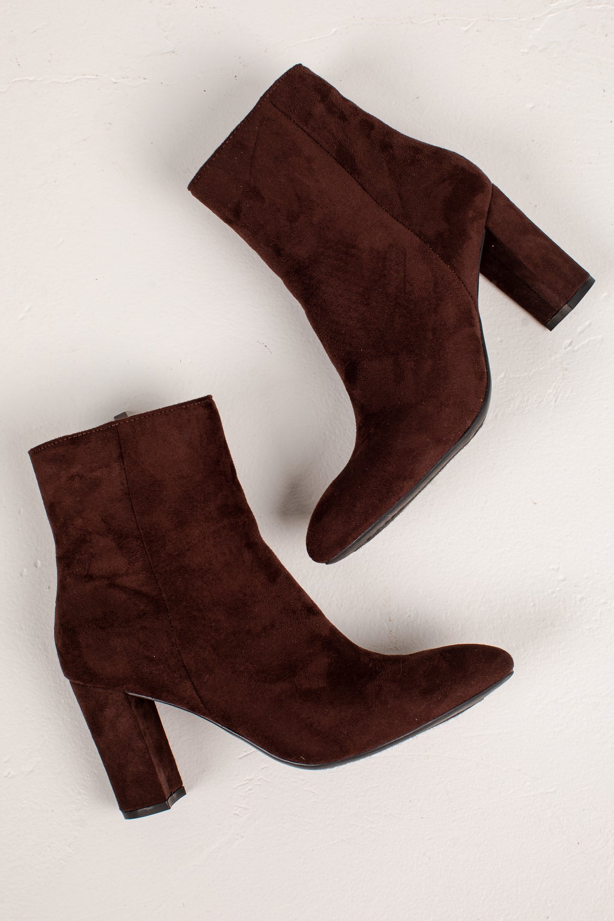 A pair of dark brown suede ankle boots with block heels, shown from different angles on a light off-white textured background.