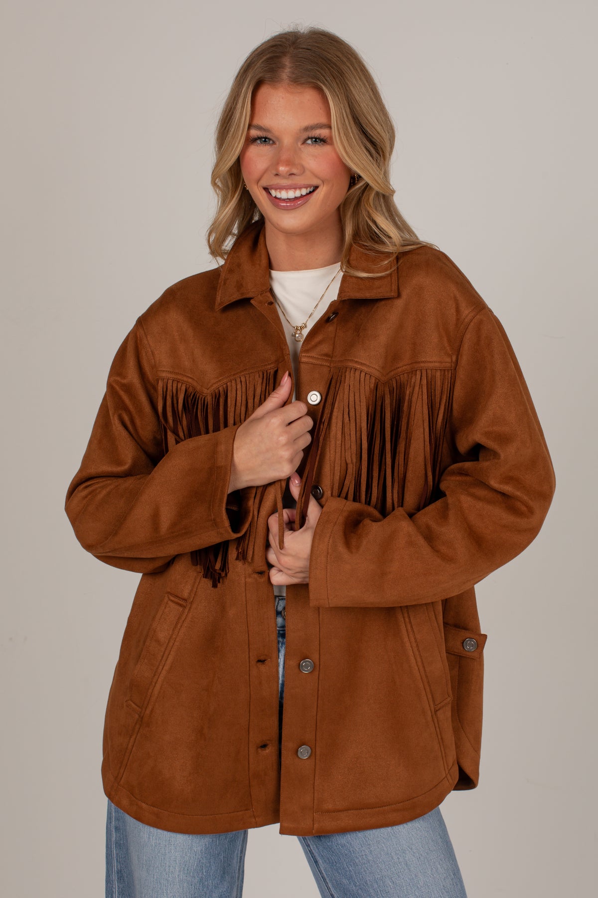 A smiling blonde woman models a brown suede-like fringed jacket over a white top and blue jeans, photographed against a plain light gray studio background.