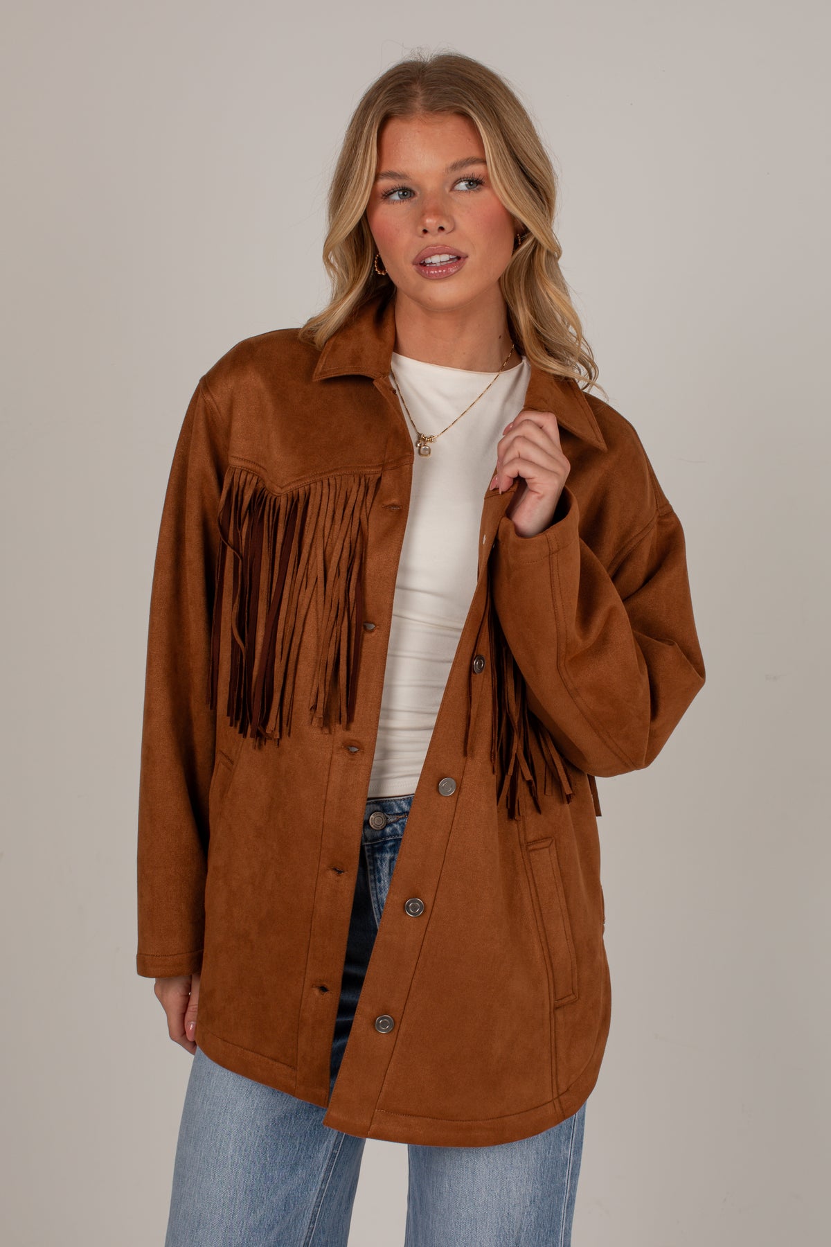 A woman modeling a brown suede fringe jacket over a white top and blue jeans, photographed against a plain light gray studio background.