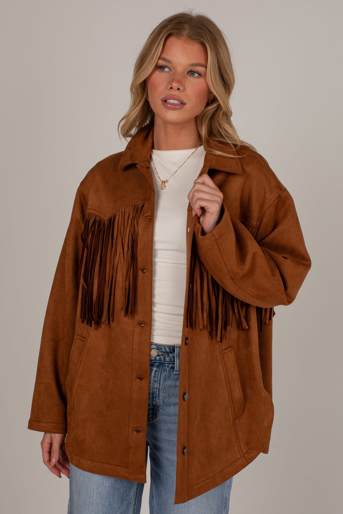 A woman modeling a brown suede fringe jacket over a white top and blue jeans, photographed against a plain light gray studio background.