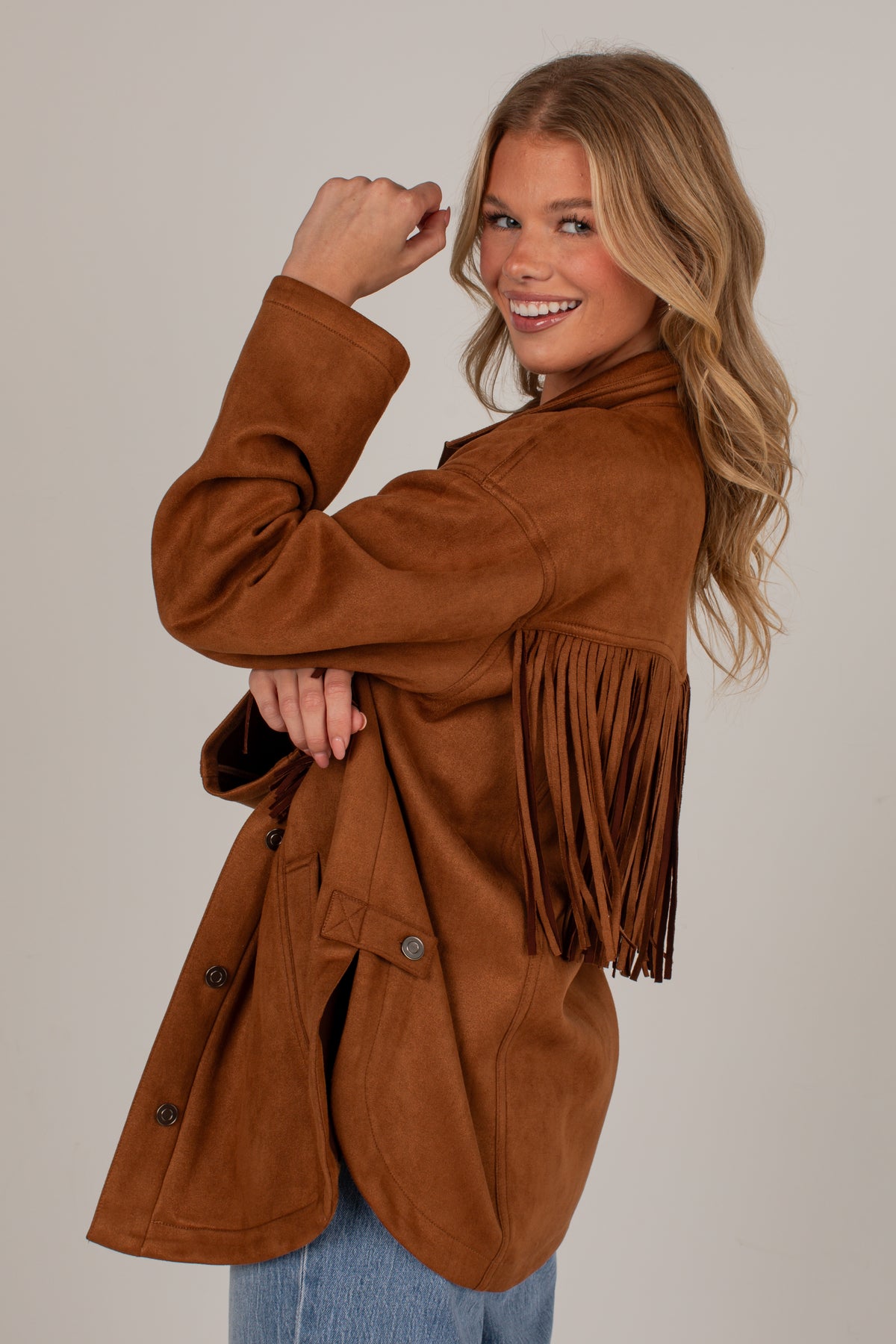 A woman modeling a brown suede fringe jacket against a solid light gray background.
