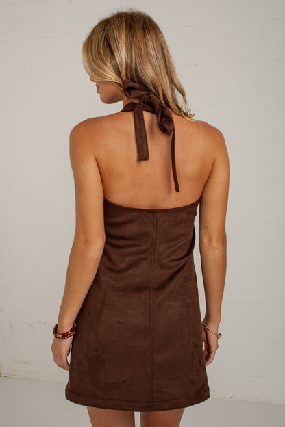A woman shown from the back wearing a brown halter-neck mini dress with a tie at the back of the neck, photographed against a plain off-white wall.