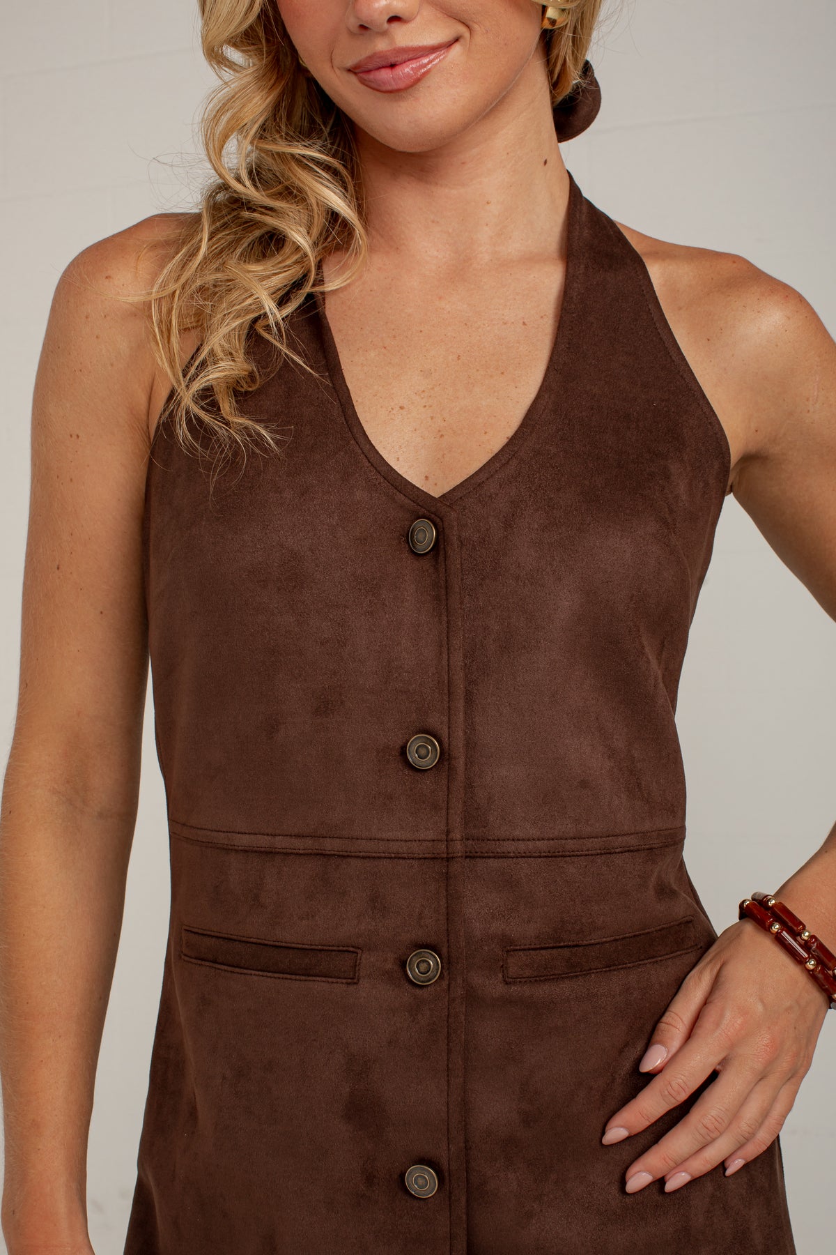 A close-up of a woman wearing a sleeveless brown faux-suede halter top with a button-front closure and front patch pockets, photographed against a plain light background.