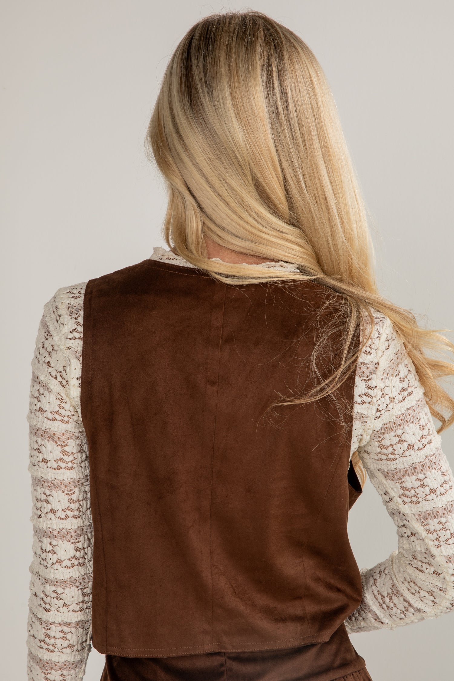 Person wearing a brown vest over a lace top against a plain background