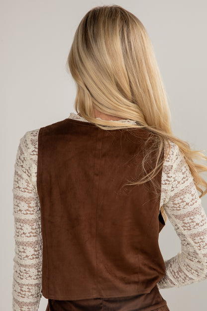 Person wearing a brown vest over a lace top against a plain background