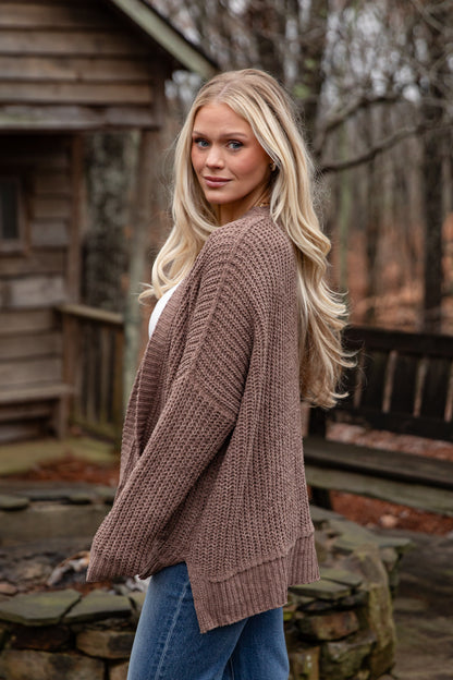 Woman wearing a brown cardigan and blue jeans standing outdoors near a wooden cabin.