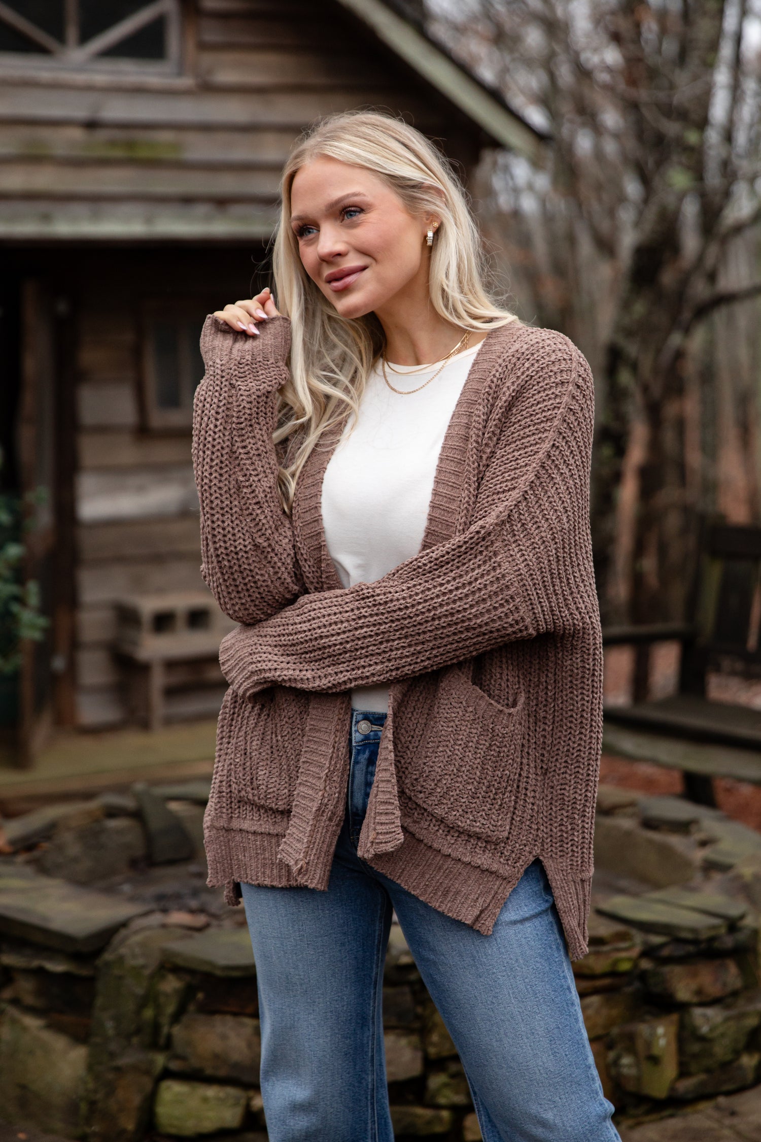 Woman wearing a brown cardigan over a white shirt and blue jeans, standing in front of a wooden cabin.