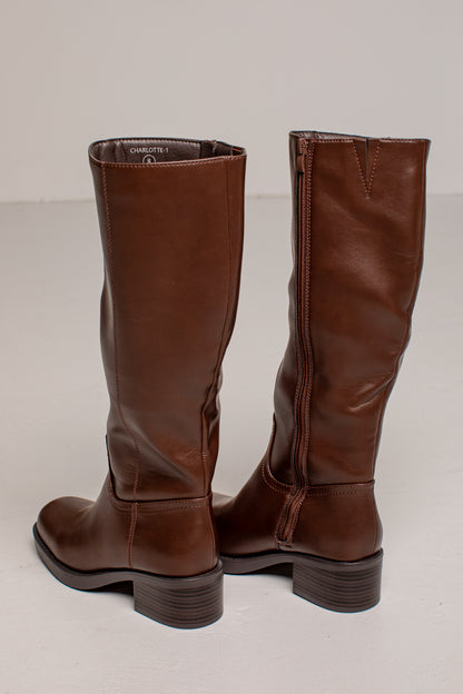 A pair of dark brown leather knee-high riding boots with low block heels, shown from the back and side against a neutral light gray background.