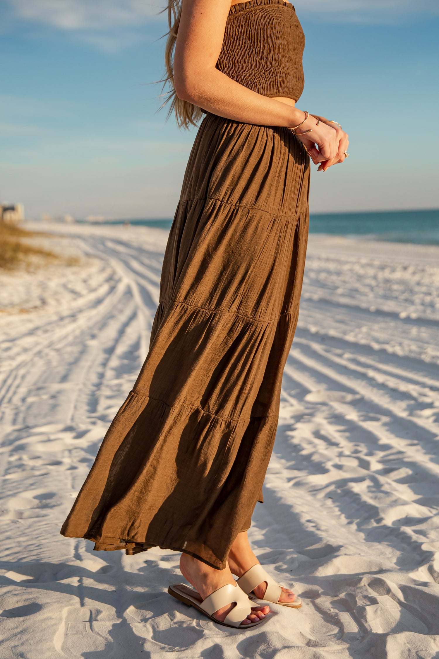 Person wearing a long brown dress on a sandy beach with ocean view
