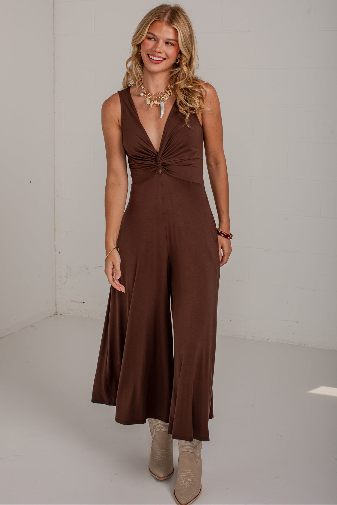 A woman modeling a sleeveless brown jumpsuit with a deep V-neckline and twisted waist detail, paired with beige ankle boots, in a bright white studio setting.