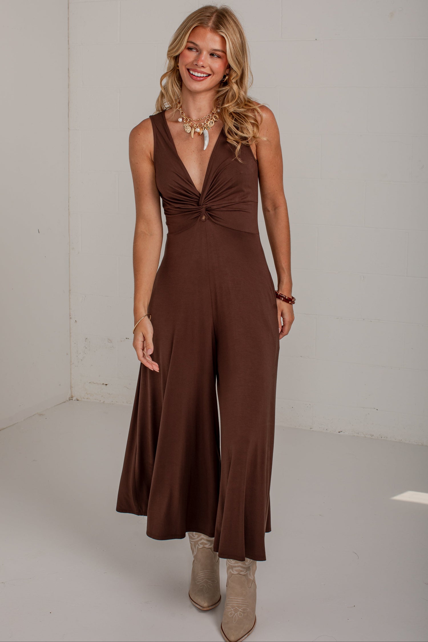 A woman modeling a sleeveless brown jumpsuit with a deep V-neckline and twisted waist detail, paired with beige ankle boots, in a bright white studio setting.