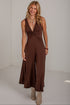 A woman modeling a sleeveless brown jumpsuit with a deep V-neckline and twisted waist detail, paired with beige ankle boots, in a bright white studio setting.