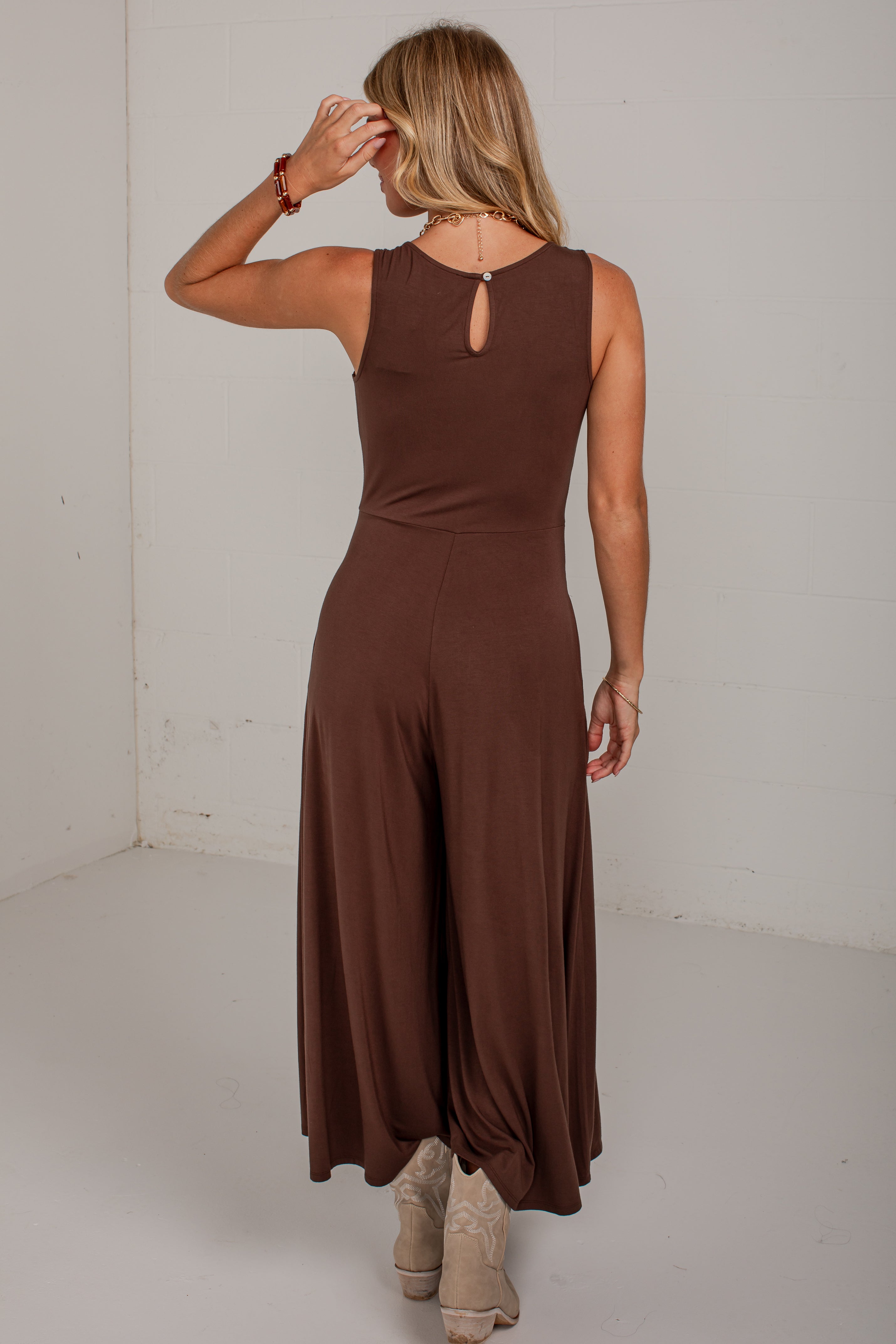 A woman wearing a sleeveless brown jumpsuit, shown from the back in a studio setting with a white wall and floor.