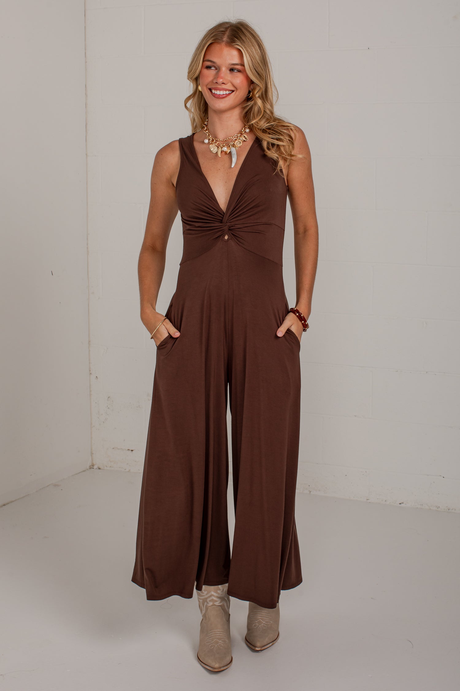 A woman modeling a sleeveless brown jumpsuit with a deep V-neckline, gathered waist detail, and wide legs, standing against a plain white studio wall.