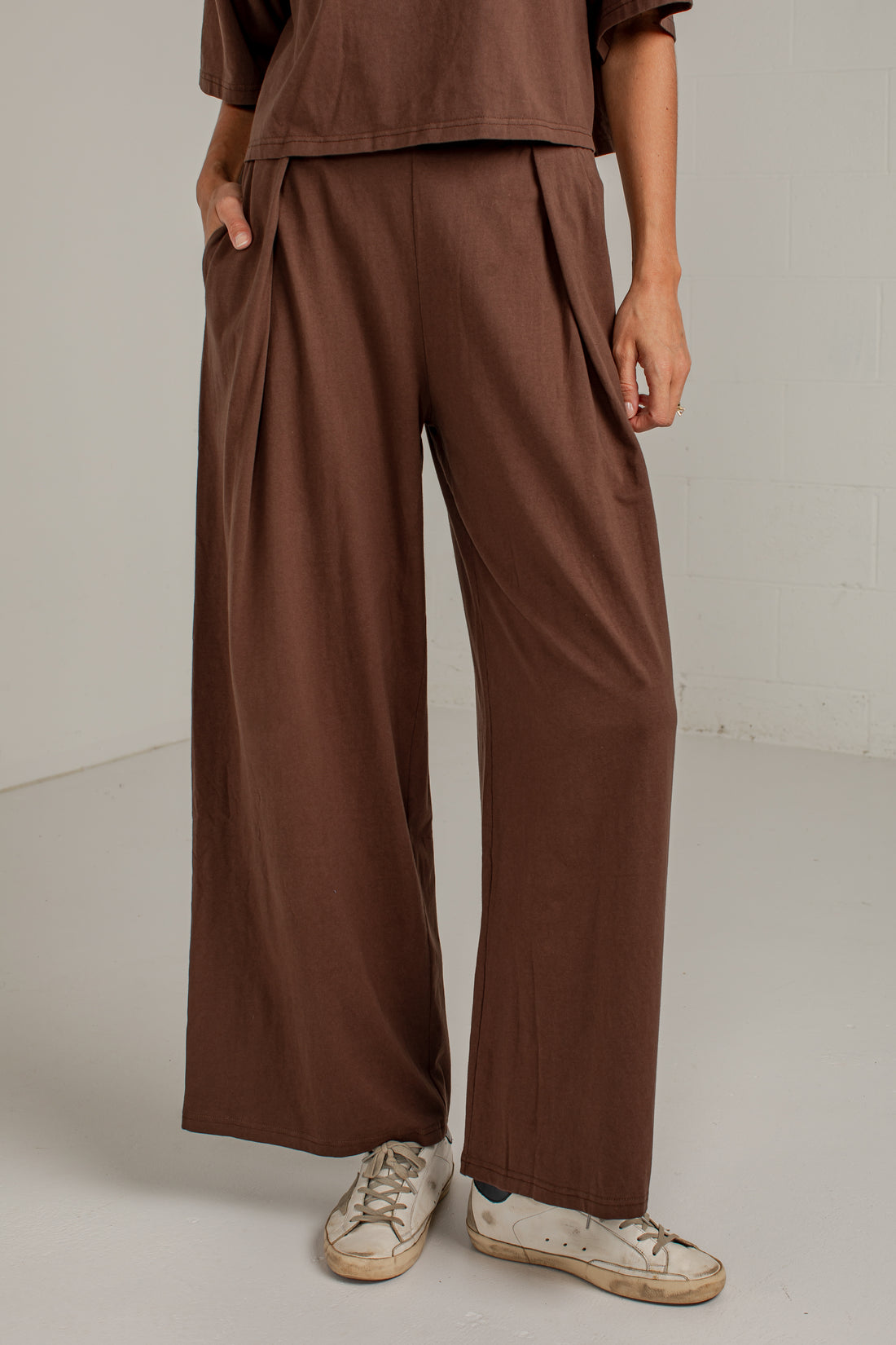 A person wearing loose, wide-leg brown pants with a matching brown top and white sneakers, standing against a plain light gray studio wall.