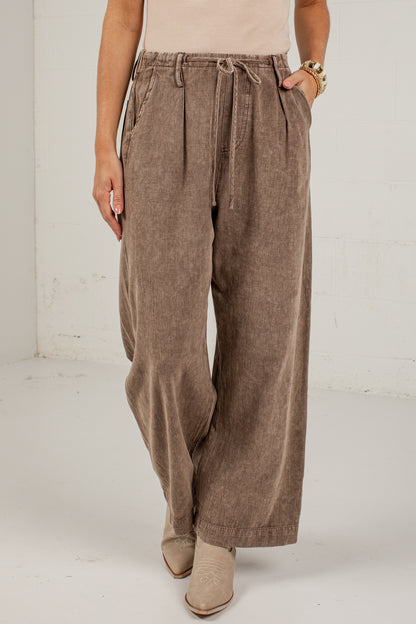 A person wearing high-waisted, wide-leg taupe corduroy pants with a drawstring waist, paired with beige ankle boots in a bright studio setting.