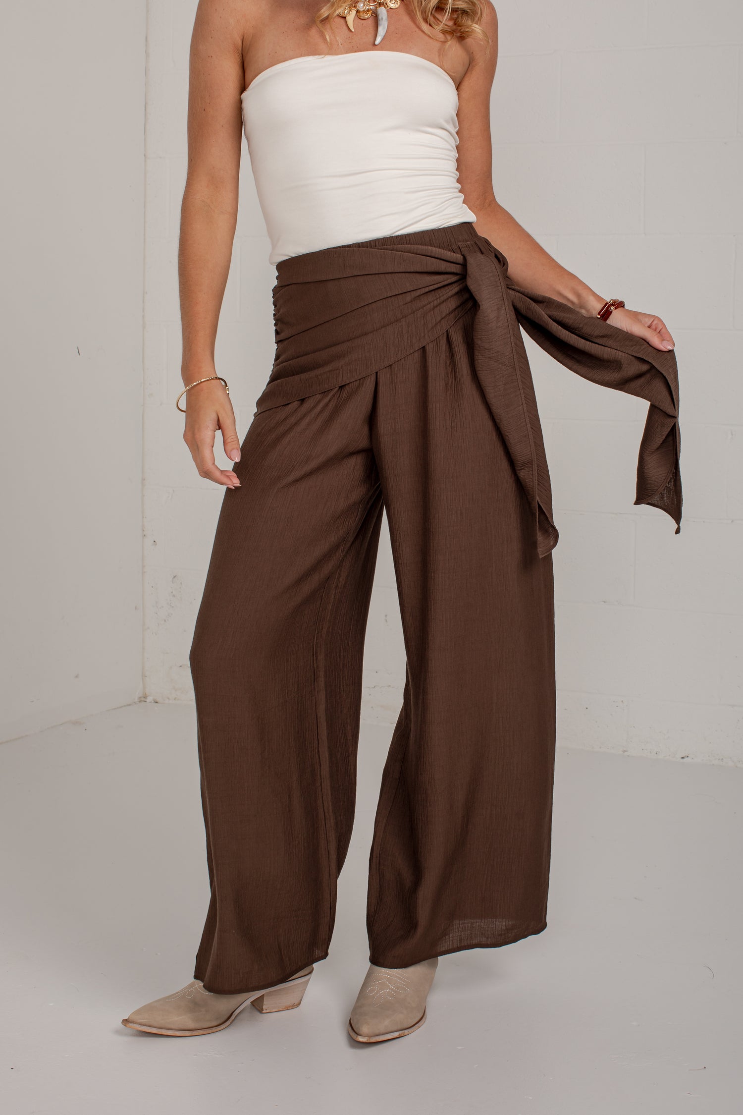 A model wearing high-waisted, wide-leg brown pants with a matching fabric sash tied at the hip, styled with a white strapless top and beige heeled shoes.