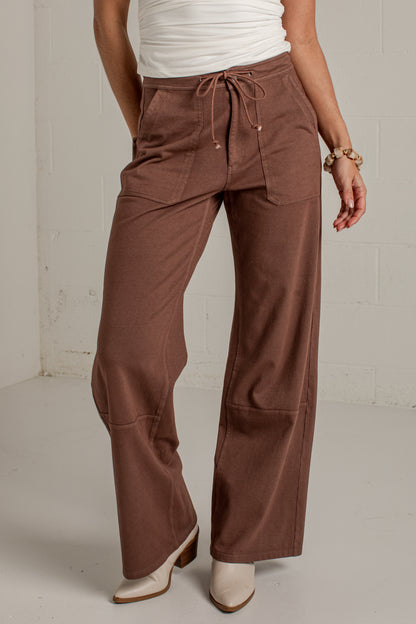 A person wearing wide-leg brown pants with a drawstring waist, paired with a white top and white heeled boots, standing against a light gray wall.
