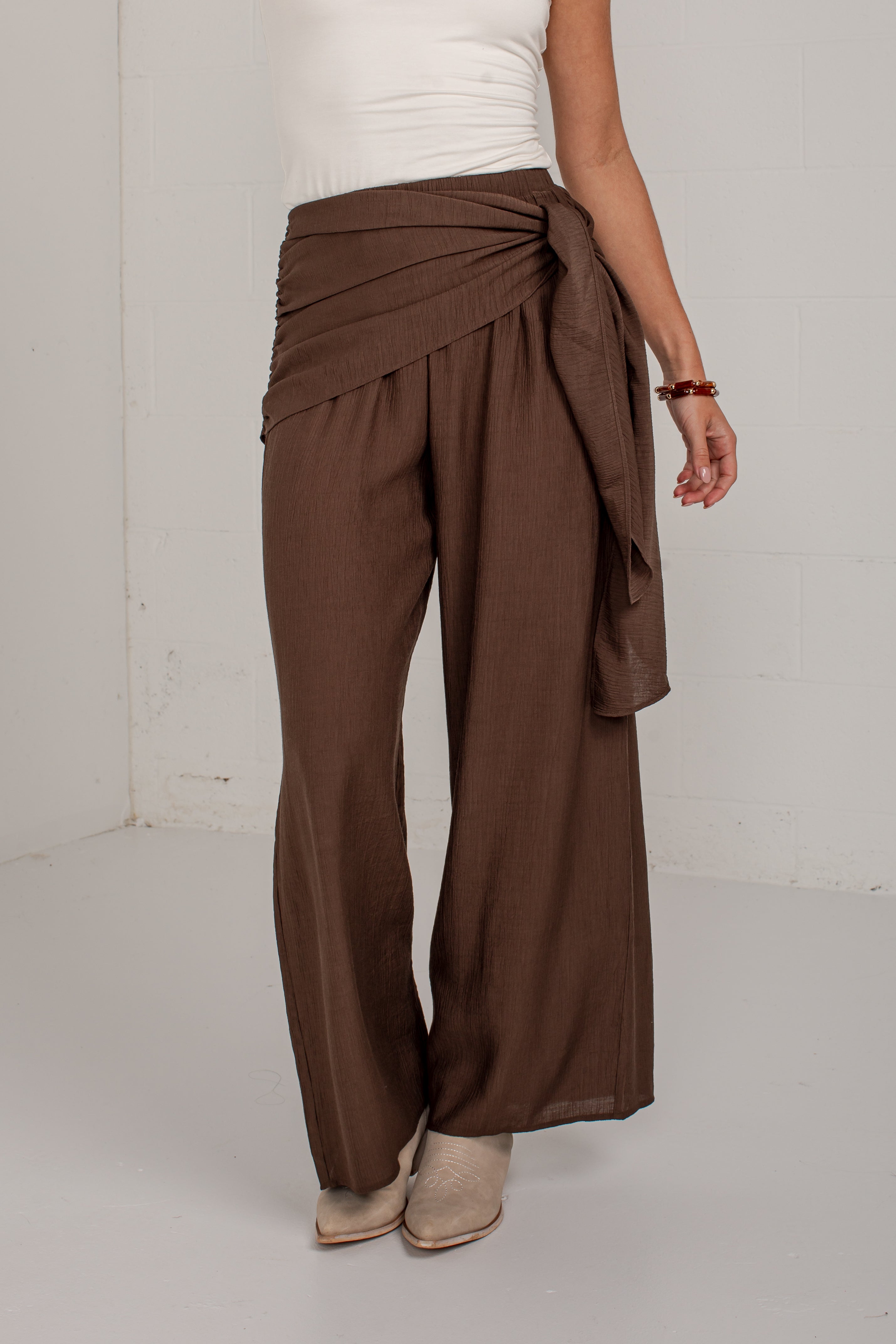 A person wearing brown wide-leg draped pants with a tied waist, paired with a white top and beige ankle boots, standing against a plain light gray wall.