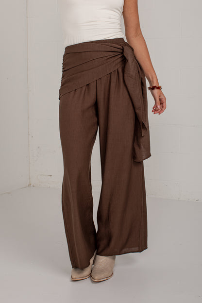 A person wearing brown wide-leg draped pants with a tied waist, paired with a white top and beige ankle boots, standing against a plain light gray wall.