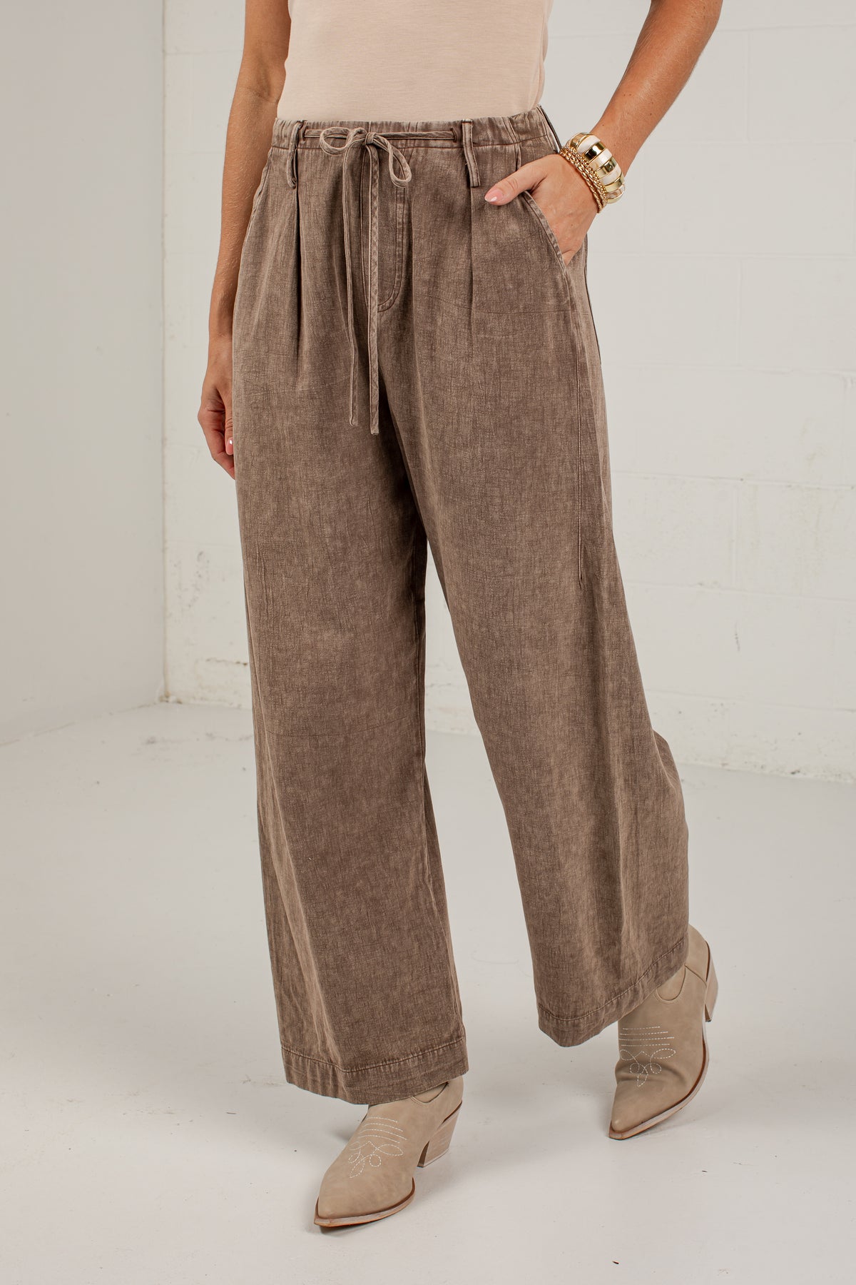 A person wearing wide-leg taupe-brown pants with a drawstring waist, paired with a light beige top and beige ankle boots, standing against a white studio wall.